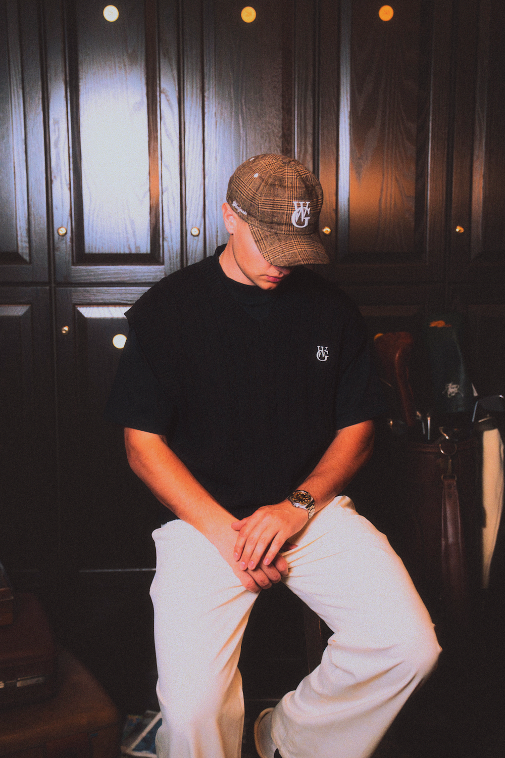 The Golfer on the clubhouse veranda in the black Perfect Trouser, tan nylon vest, and plaid wool cap